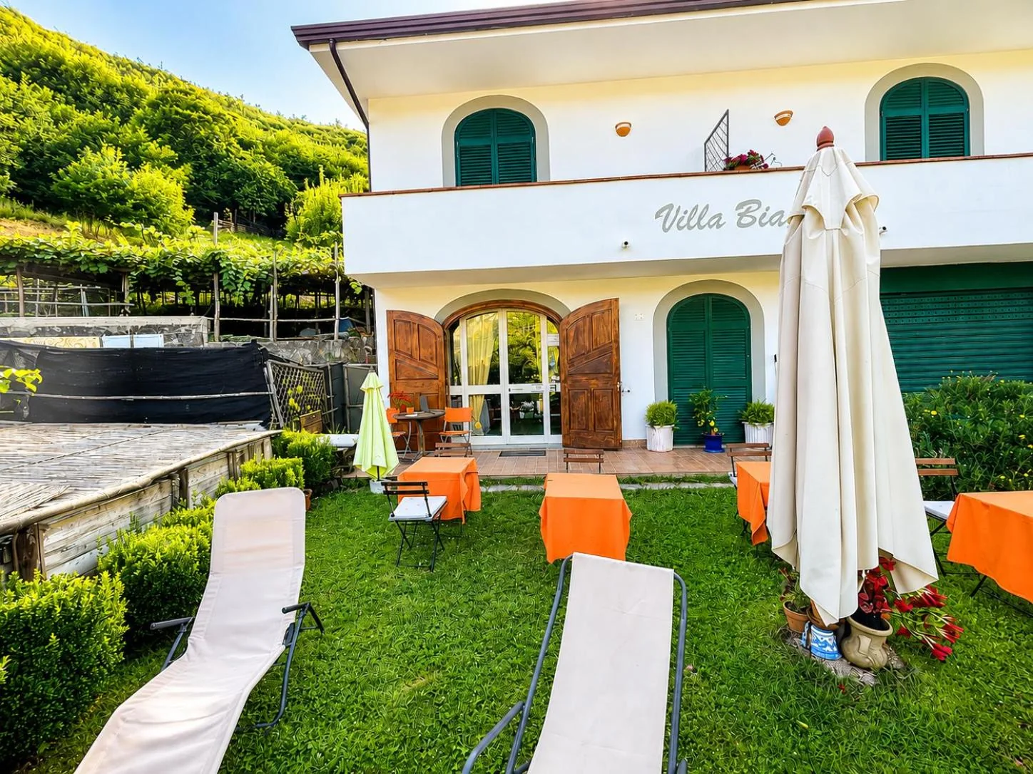 Villa Bianca vista dal giardino: la facciata bianca con le imposte verdi, i lettini sul prato e il vigneto terrazzato alle spalle